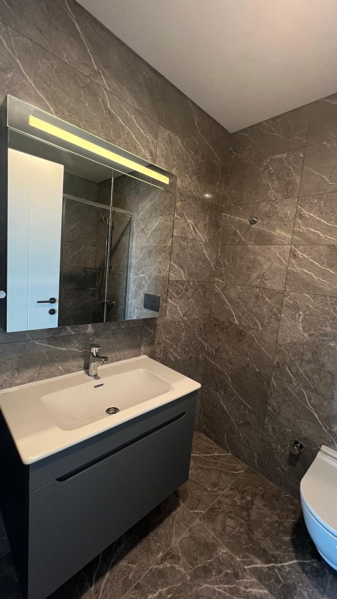 En-suite bathroom with marble-look tiles, a vanity unit, an illuminated mirror cabinet and a wall-hung toilet