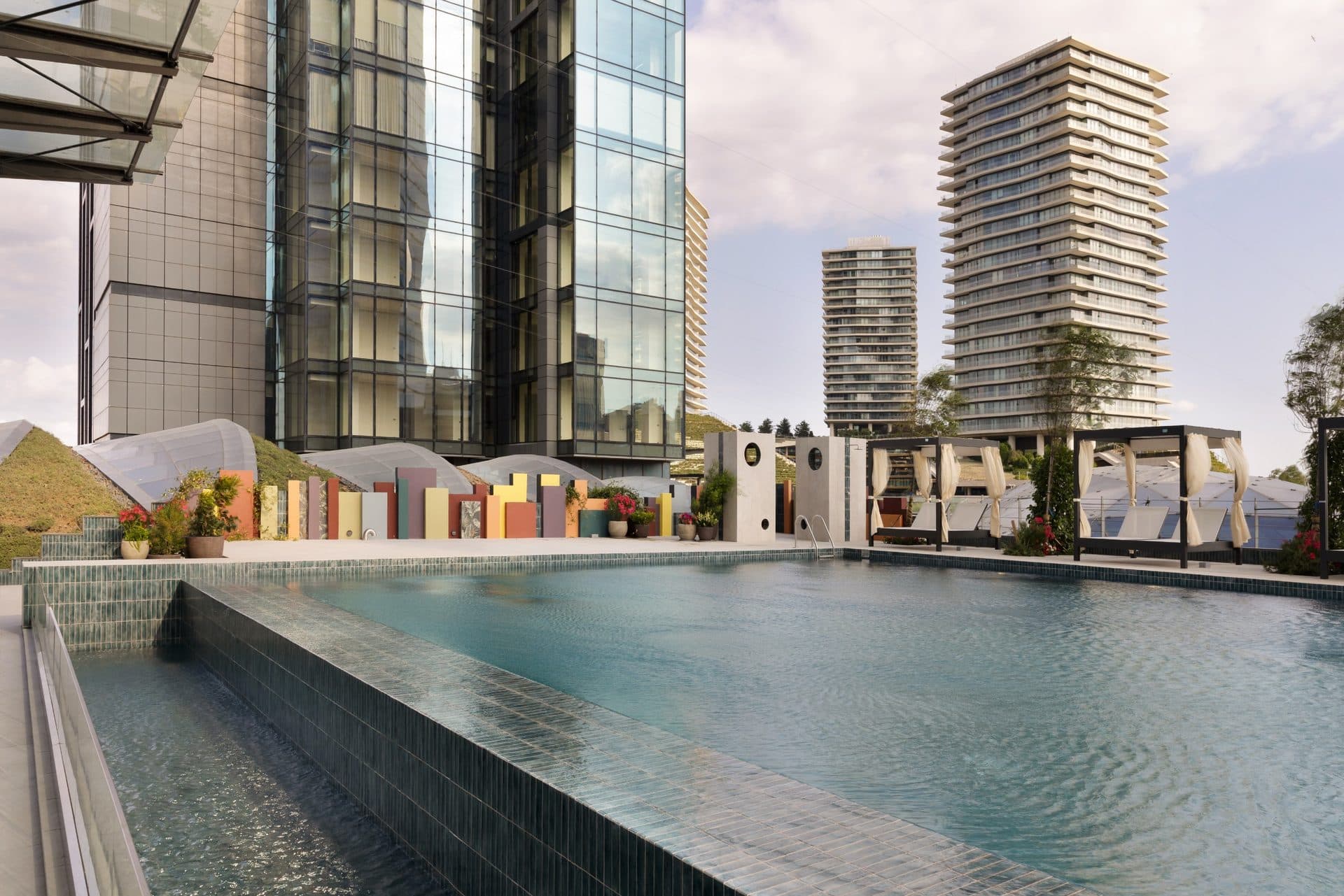 Outdoor swimming pool with cabanas on the podium terrace, beside the glass facade of one of the towers