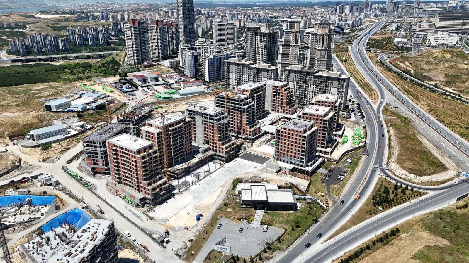 Drone photo from June 2026 with the sales office and its car park in front of the blocks and the boulevard curving past on the right