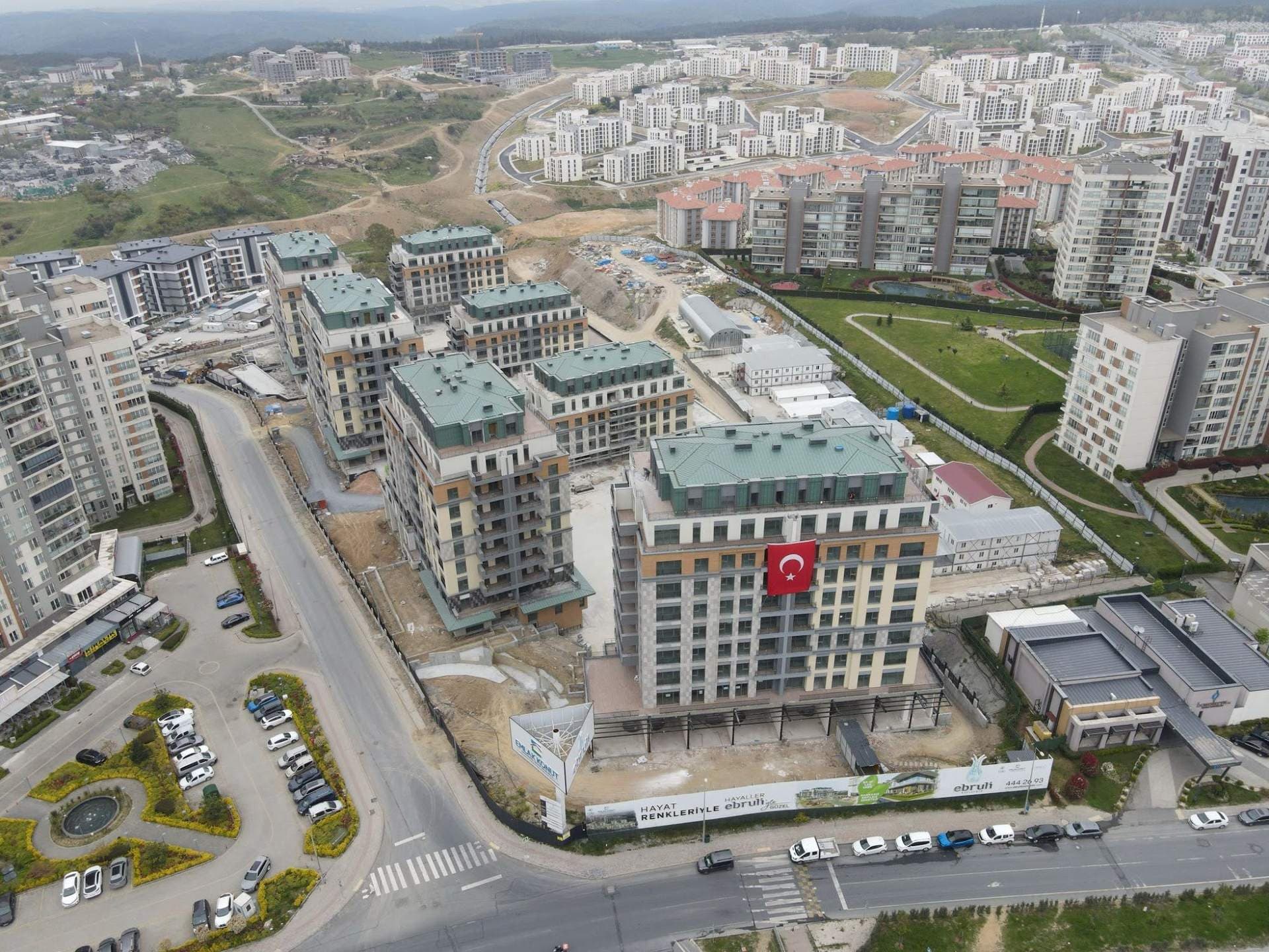 Drone photo from May 2025 showing the blocks with their green roofs in place and the corner block flying a Turkish flag, while site works continue around them