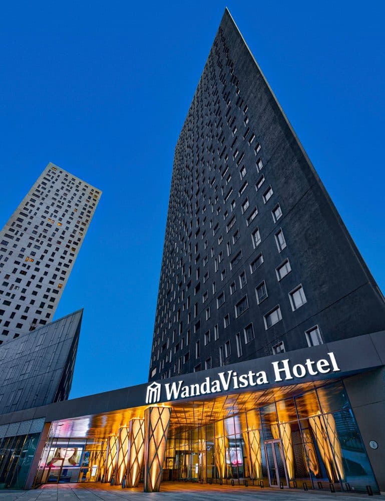 The dark grey hotel tower seen from below against a deep blue evening sky, with the lit WandaVista Hotel entrance canopy at its base and the white G Yoo tower rising on the left