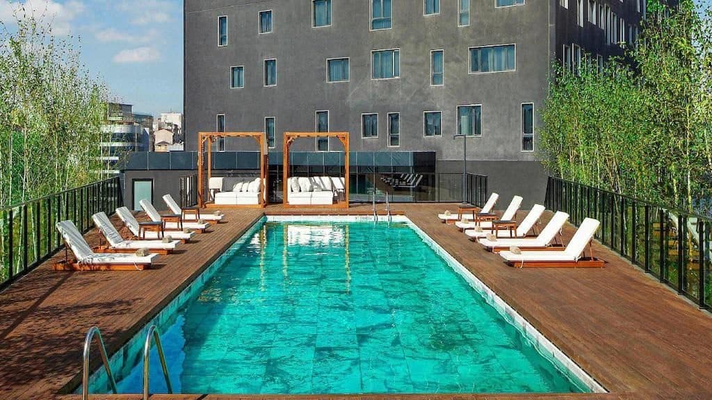 Outdoor swimming pool on the podium terrace, lined with white sun loungers and two canopied daybeds on a timber deck, with the hotel's lower floors behind