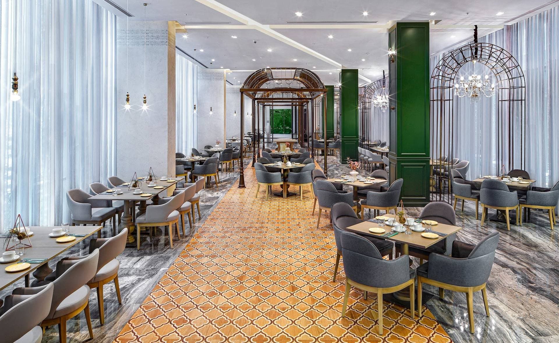 All-day restaurant with a patterned parquet floor, green columns, grey upholstered chairs and laid tables beside tall curtained windows