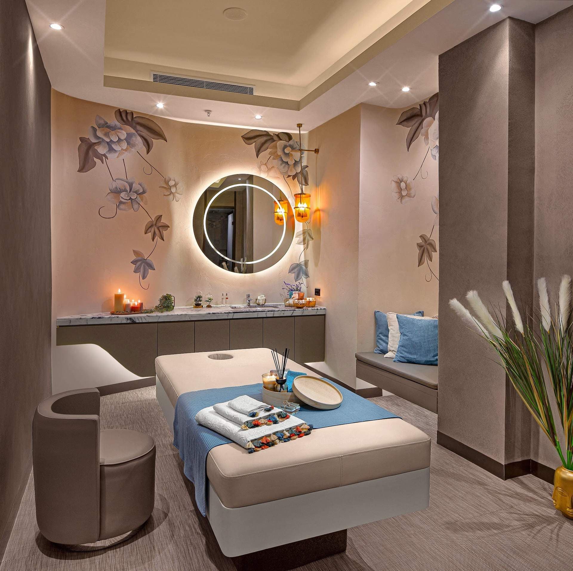 Spa treatment room with a massage bed, a round backlit mirror, floral wall art and a lounge chair