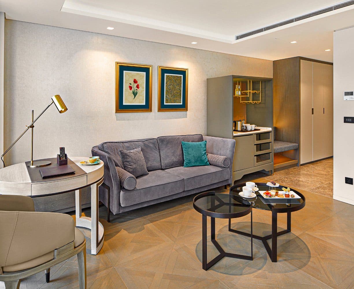 Furnished living area with a grey velvet sofa, round glass coffee tables, a writing desk with a brass lamp and a built-in minibar unit on a herringbone parquet floor