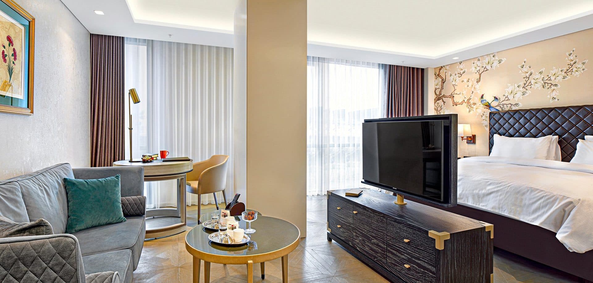 Open-plan suite with a double bed behind a low TV console, a sofa and a small dining table by the window, and floral wallpaper behind the bed