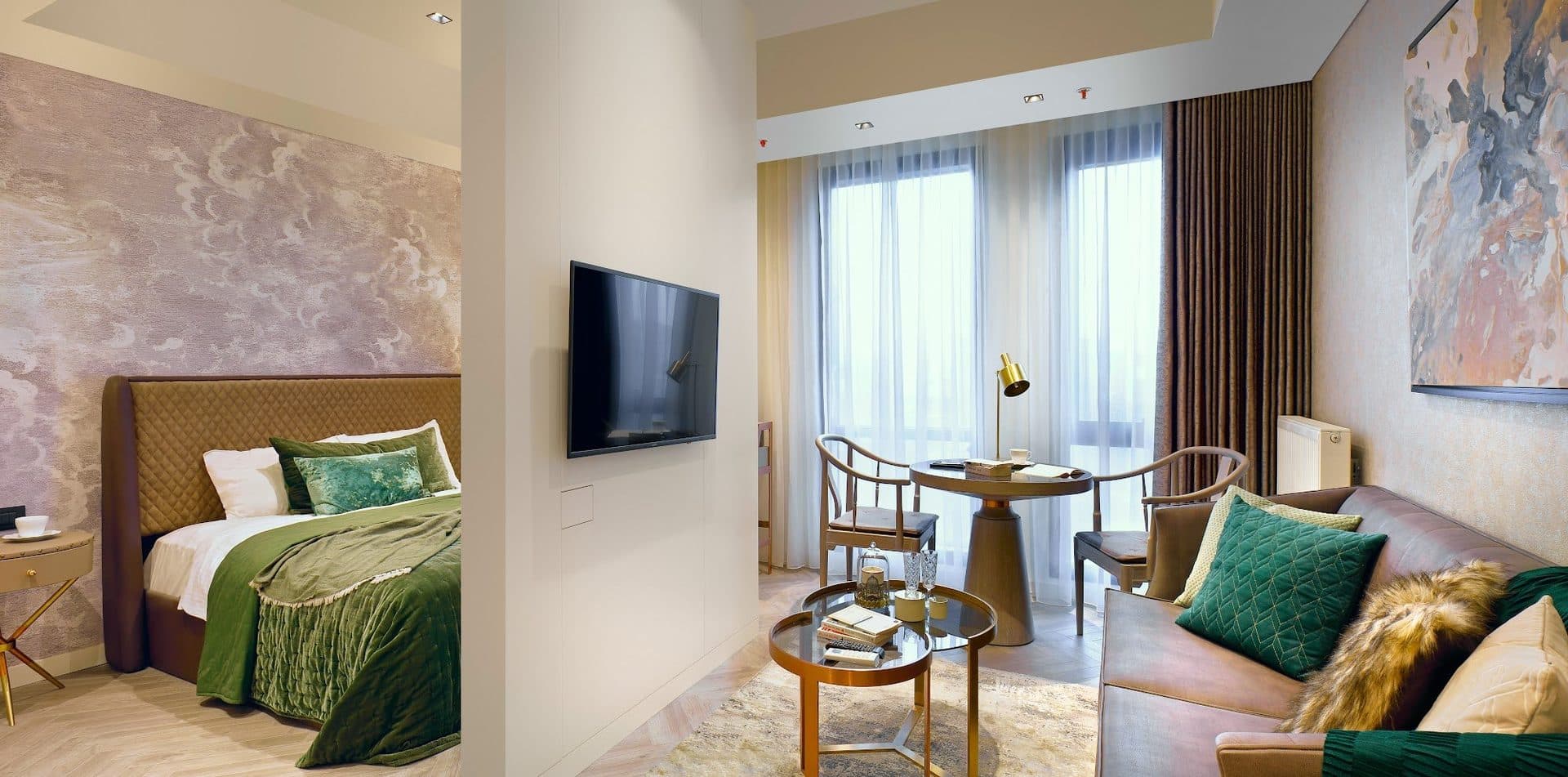 Residence with a bedroom beside a living area: a bed with a green throw, a wall-mounted TV, a small round table with two chairs and a leather sofa by the windows