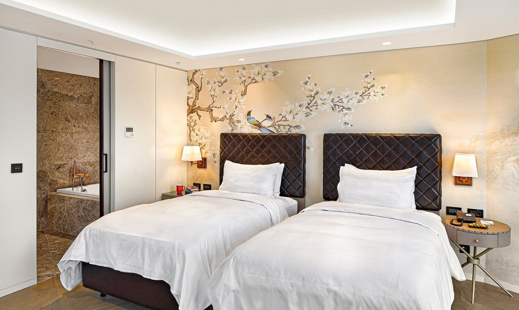 Twin bedroom with quilted headboards, bedside lamps and floral wallpaper, the marble bathroom visible through a sliding door