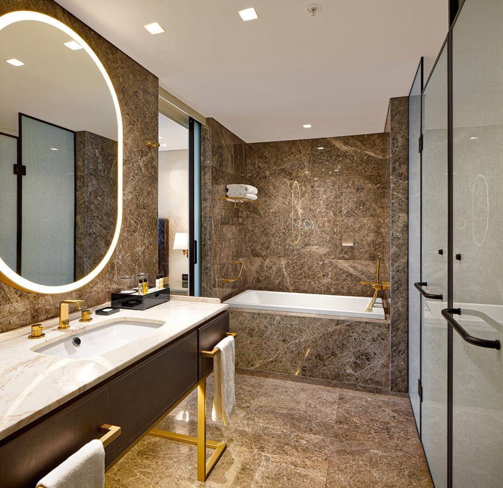 Bathroom in brown marble with a bathtub, a glass-walled shower, a backlit oval mirror and brass fittings