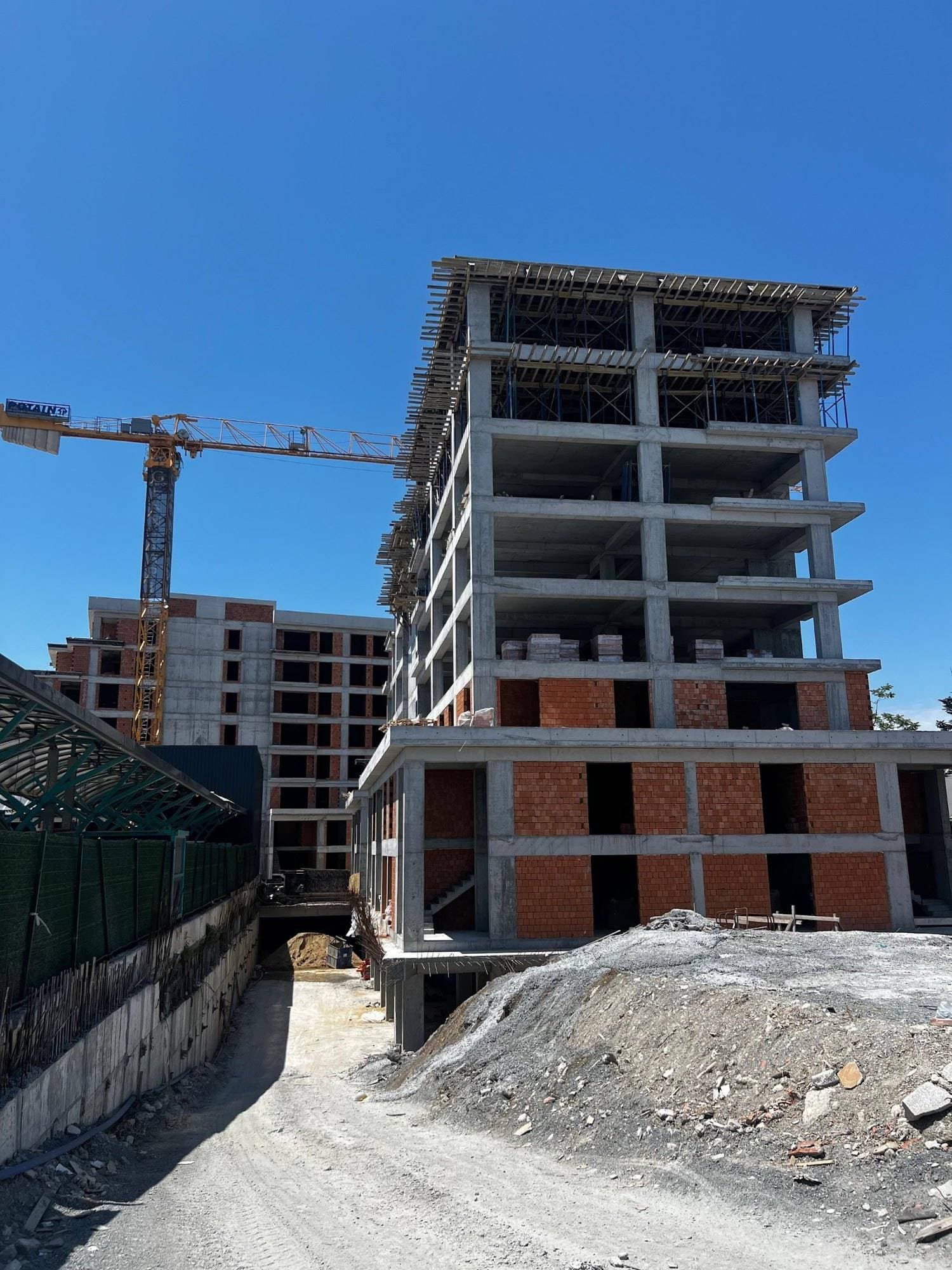 Construction site in June 2026: the concrete frame of one block with formwork on its top floor, and a block with finished brick walls and a tower crane behind