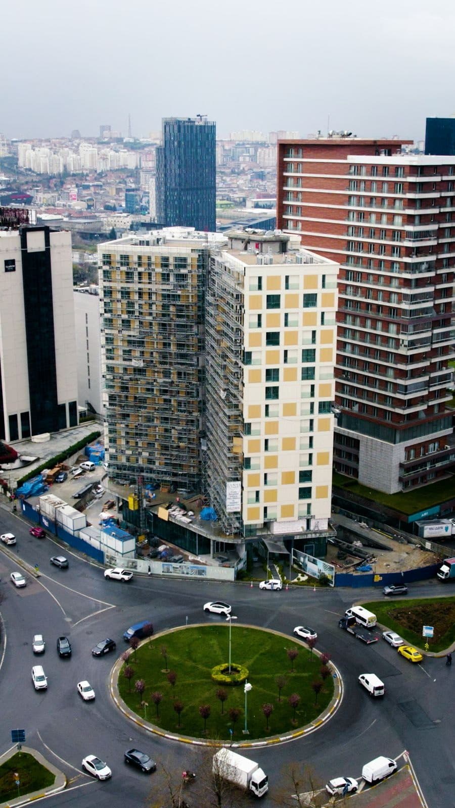 Drone photo of Lania Residence under construction in spring 2026, one wing still in scaffolding and the other with its facade panels in place, beside a roundabout
