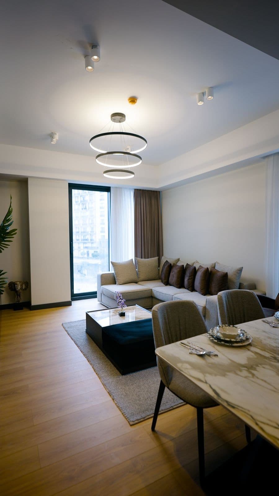 Living room of the furnished sample apartment, with a corner sofa, marble-topped tables and a full-height window