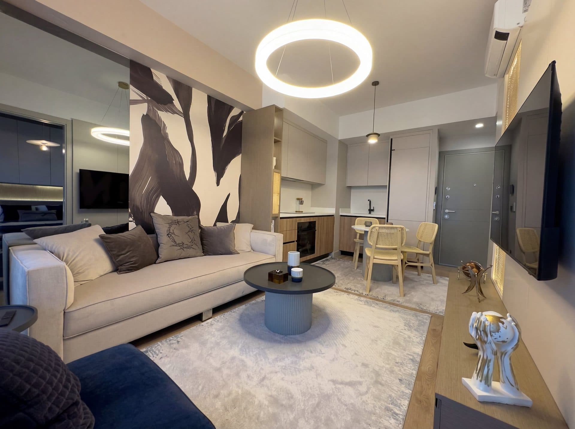 Photo of the furnished 1+1 show apartment: living room with a sofa, round coffee tables, a wall-mounted TV and the open kitchen behind