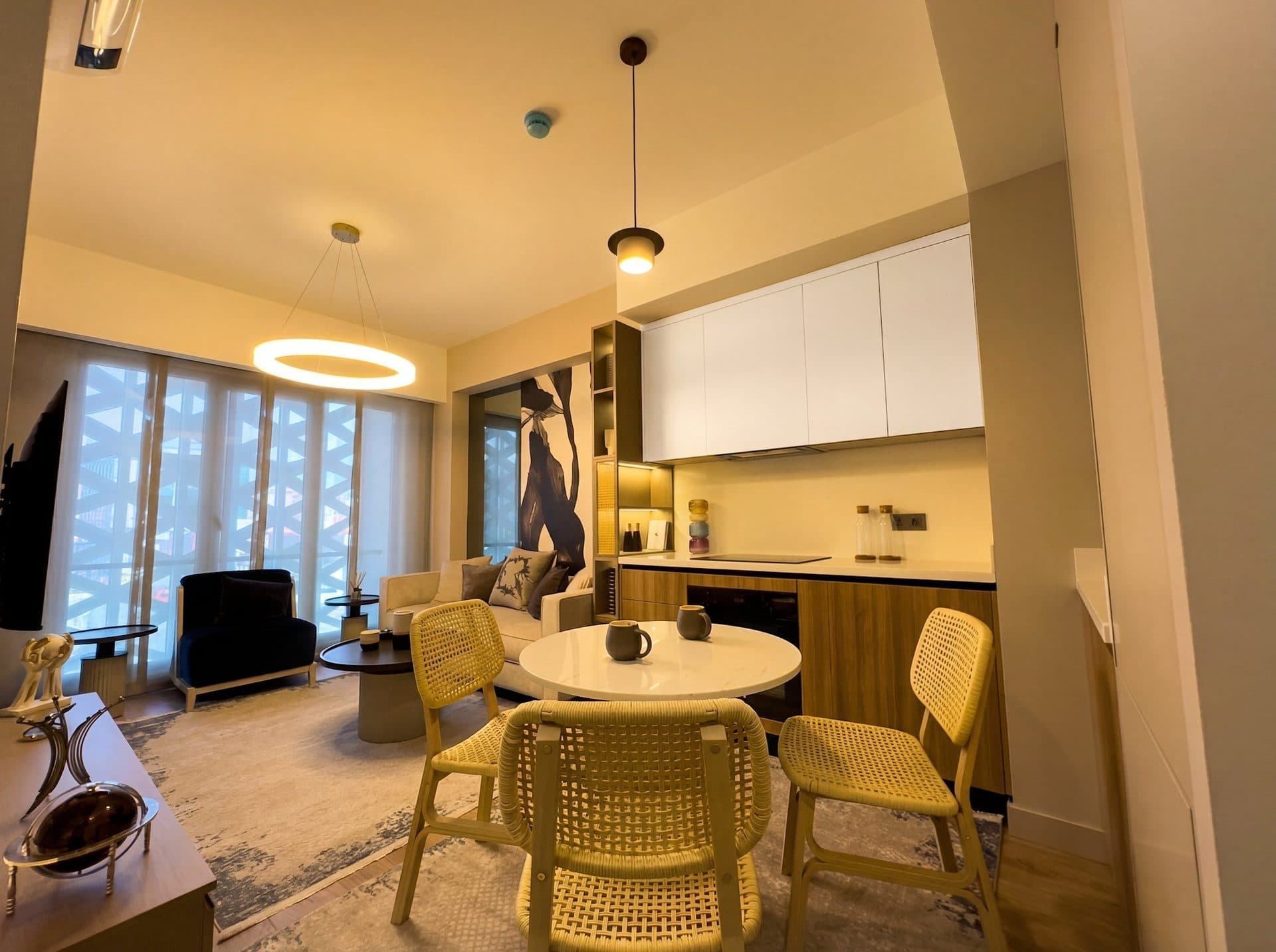 Photo of the furnished 1+1 show apartment: dining corner with a round table and wicker chairs beside the open kitchen with white wall cabinets