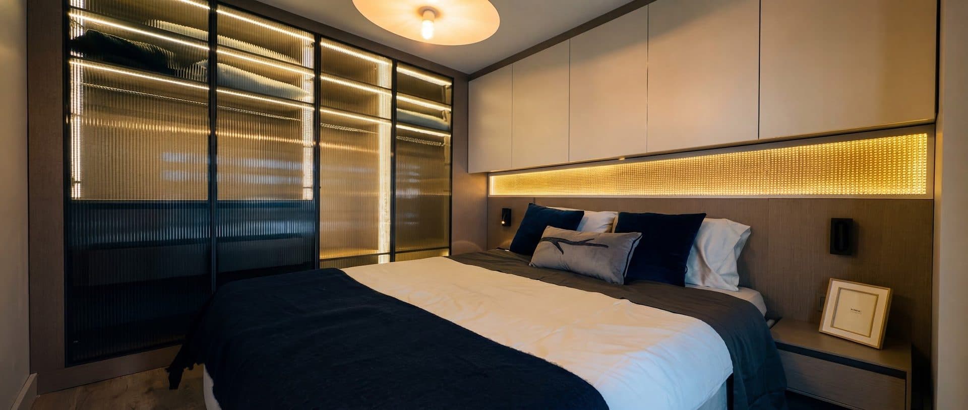 Photo of the furnished 1+1 show apartment: bedroom with a double bed, a lit shelf above the headboard and a glass-fronted wardrobe