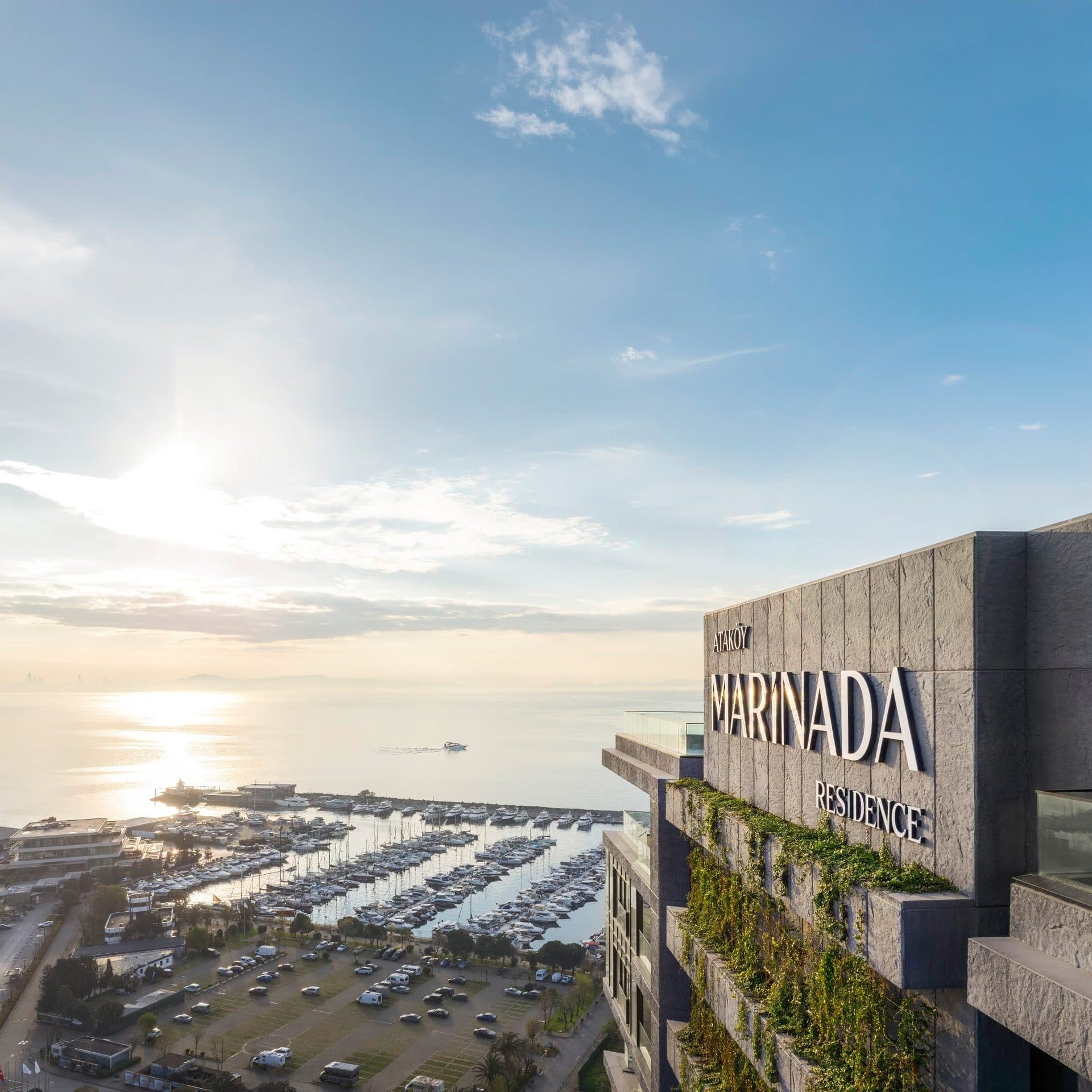 Stone-clad crown of the tower with the Marinada Residence lettering and planted terraces, with the marina below and the sea in the morning sun