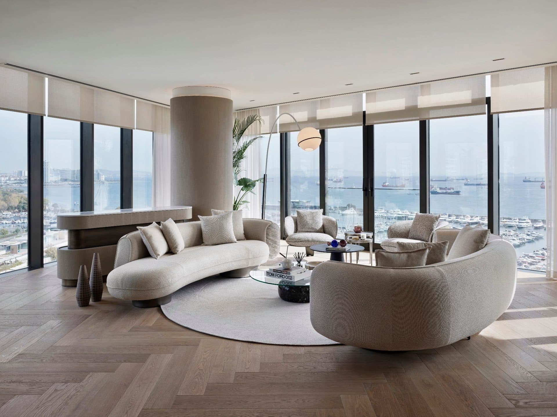 Living room of the furnished sample apartment with curved sofas and floor-to-ceiling windows facing the marina and the sea
