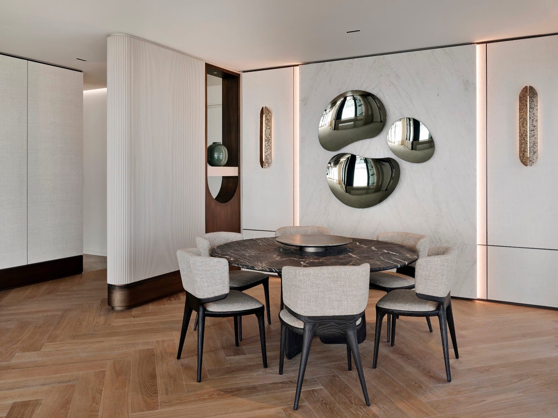Dining area with a round marble table, upholstered chairs, herringbone parquet and sculpted wall mirrors