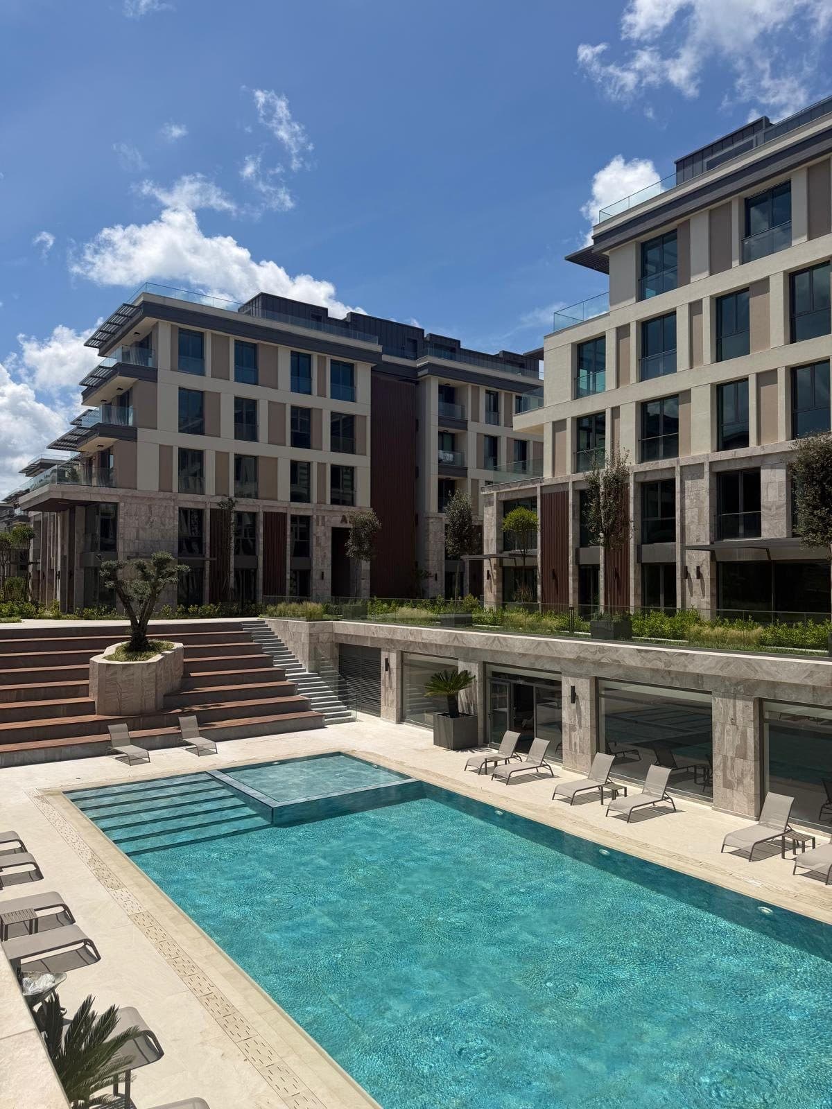 Photo of the outdoor swimming pool with sun loungers and stepped seating, in front of finished stone-clad blocks with balconies