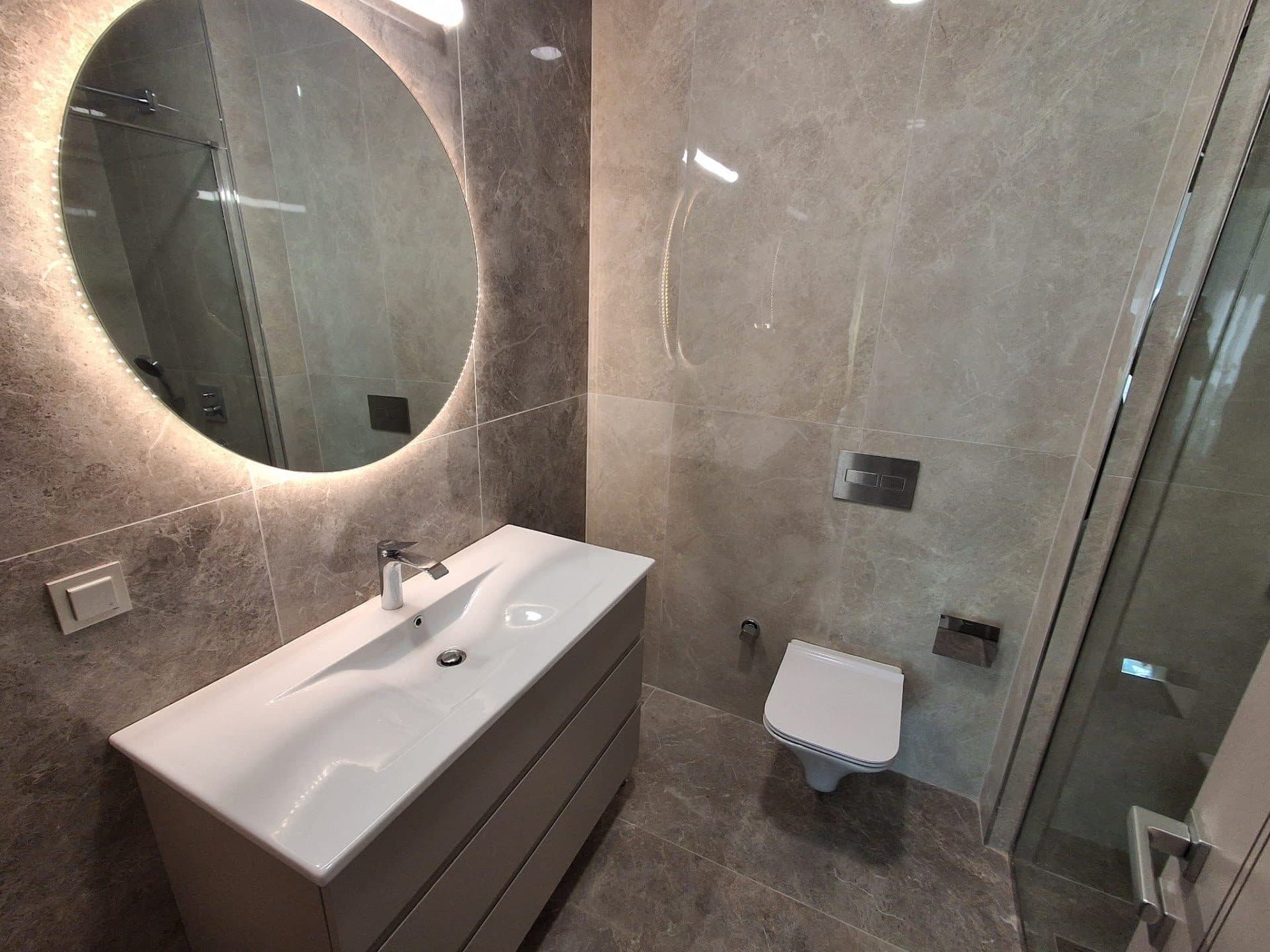Bathroom with stone-look tiles, a backlit round mirror, a wall-hung toilet and a glass shower enclosure